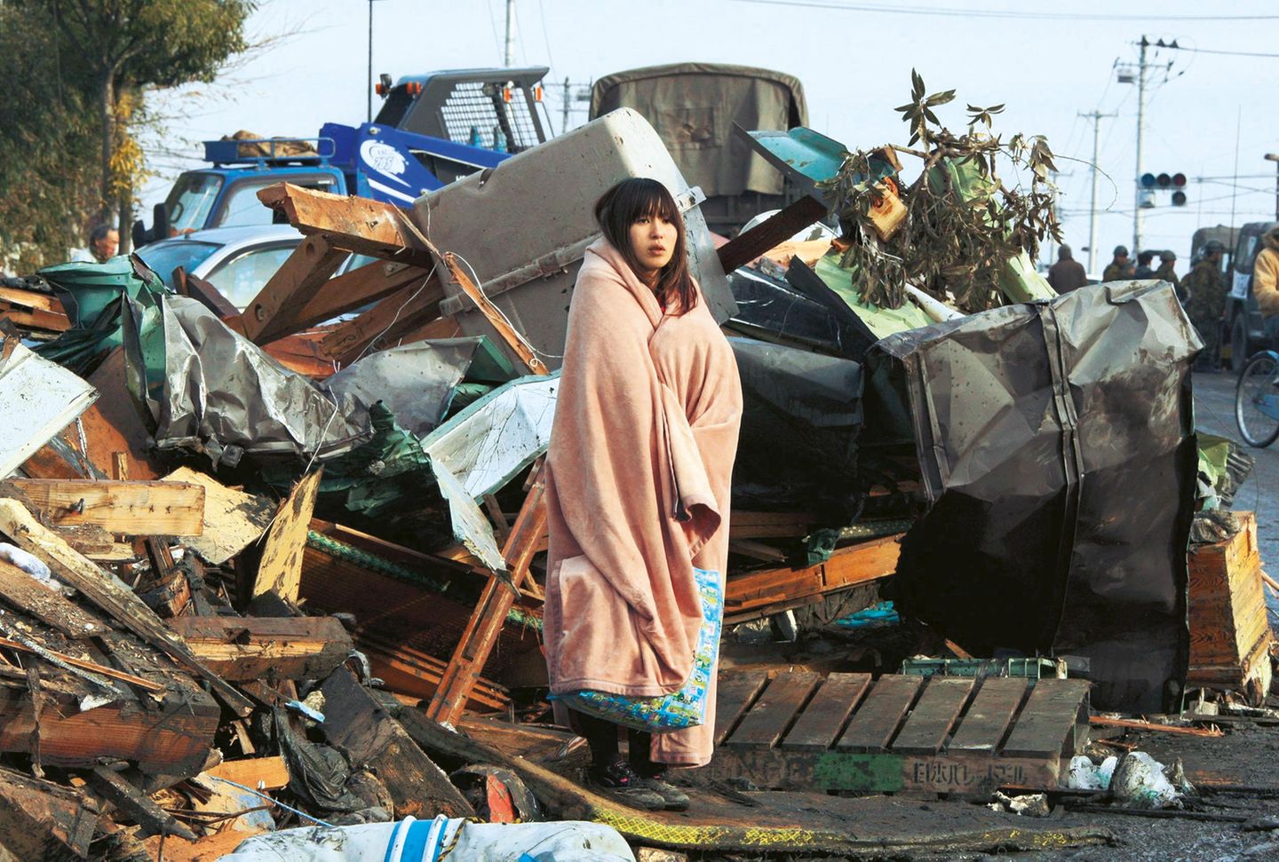 Eine junge Frau steht in eine Decke gewickelt inmitten von Trümmern.