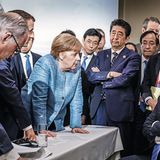 Angela Merkel lehnt sich neben anderen Regierungschefs auf einen Tisch, vor ihr verschränkt Donald Trump sitzend die Arme