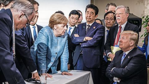 Angela Merkel lehnt sich neben anderen Regierungschefs auf einen Tisch, vor ihr verschränkt Donald Trump sitzend die Arme
