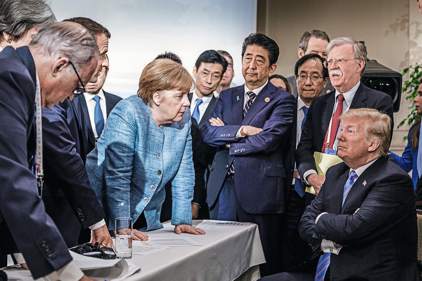 2018 – Kanada Angela Merkel lehnt sich neben anderen Regierungschefs auf einen Tisch, vor ihr verschränkt Donald Trump sitzend die Arme
