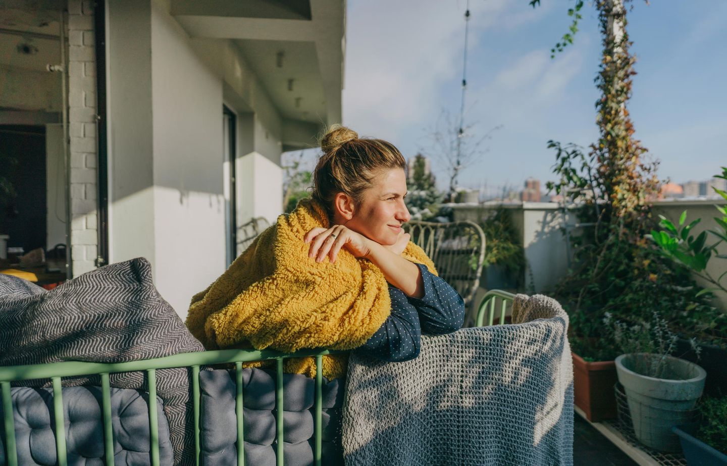Auszeit Zuhause Eine Frau sitzt auf dem Balkon in eine Decke gewickelt und genießt die Sonne.