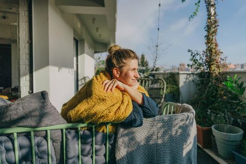Eine Frau sitzt auf dem Balkon in eine Decke gewickelt und genießt die Sonne.