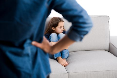Eine erwachsene Person steht mit dem Rücken zur Kamera vor einem Sofa, auf dem ein zusammengekauertes Kind sitzt