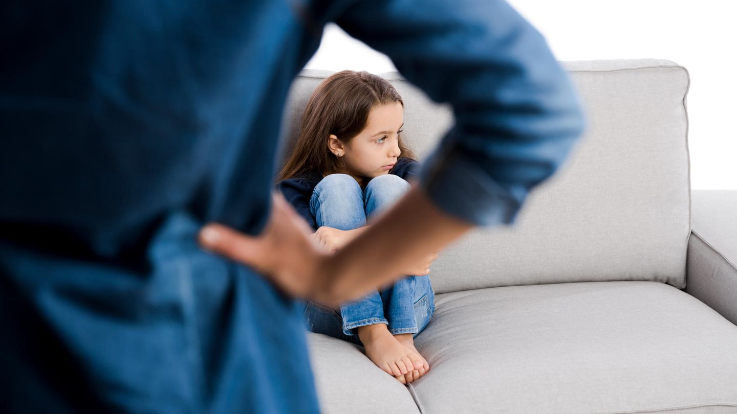 Eine erwachsene Person steht mit dem Rücken zur Kamera vor einem Sofa, auf dem ein zusammengekauertes Kind sitzt