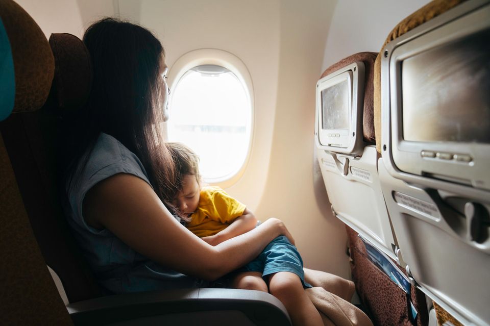 Reisetrends Eine Frau sitzt mit ihrem schlafenden Kind auf dem Schoß in einem Flugzeug und schaut aus dem Fenster.