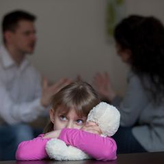 Scheidung: Ein kleines Mädchen umarmt ihr Kuscheltier, während sich ihre Eltern im Hintergrund streiten