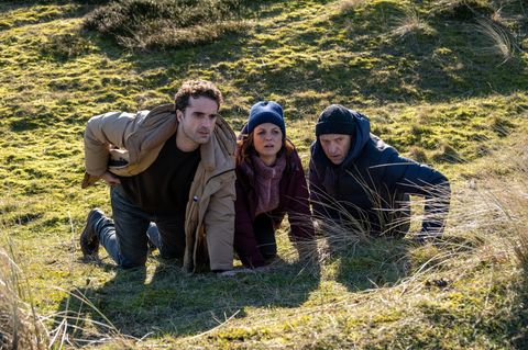 Carl Sievers (Peter Heinrich Brix, r.), Hinnerk Feldmann (Oliver Wnuk) und Ina Behrendsen (Julia Brendler) suchen Schutz hinter einem Hügel in einer Szene des Films "Nord Nord Mord - Sievers und die fünf Fragezeichen"