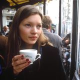 Kate Moss mit roten Nägeln und einer Tasse in der Hand. Sie sitzt in einem Café und schaut aus dem Fenster