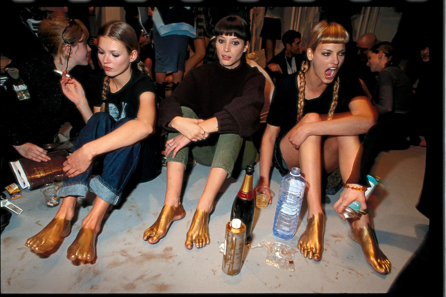 Kate Moss, Christy Turlington und Linda Evangelista sitzen nebeneinander auf dem Boden. Ihre Füße sind Gold eingesprüht.