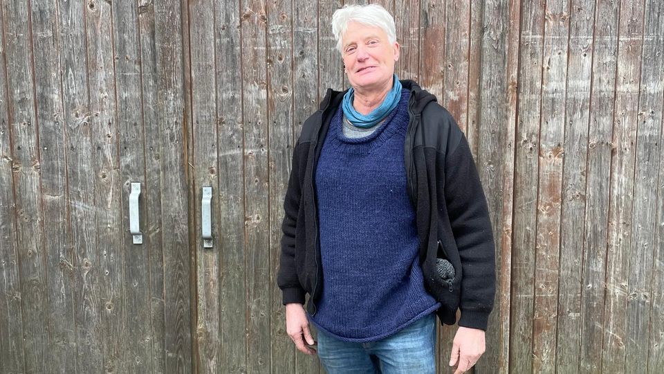 Ein Mann mit weißen Haaren und Fleecejacke steht vor einem Scheunentor. Ein Mann mit weißen Haaren und Fleecejacke steht vor einem Scheunentor.