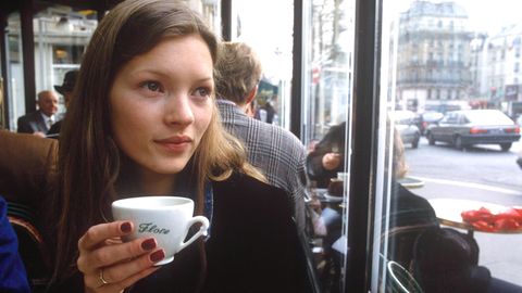 Kate Moss mit roten Nägeln und einer Tasse in der Hand. Sie sitzt in einem Café und schaut aus dem Fenster
