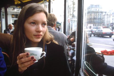Kate Moss mit roten Nägeln und einer Tasse in der Hand. Sie sitzt in einem Café und schaut aus dem Fenster