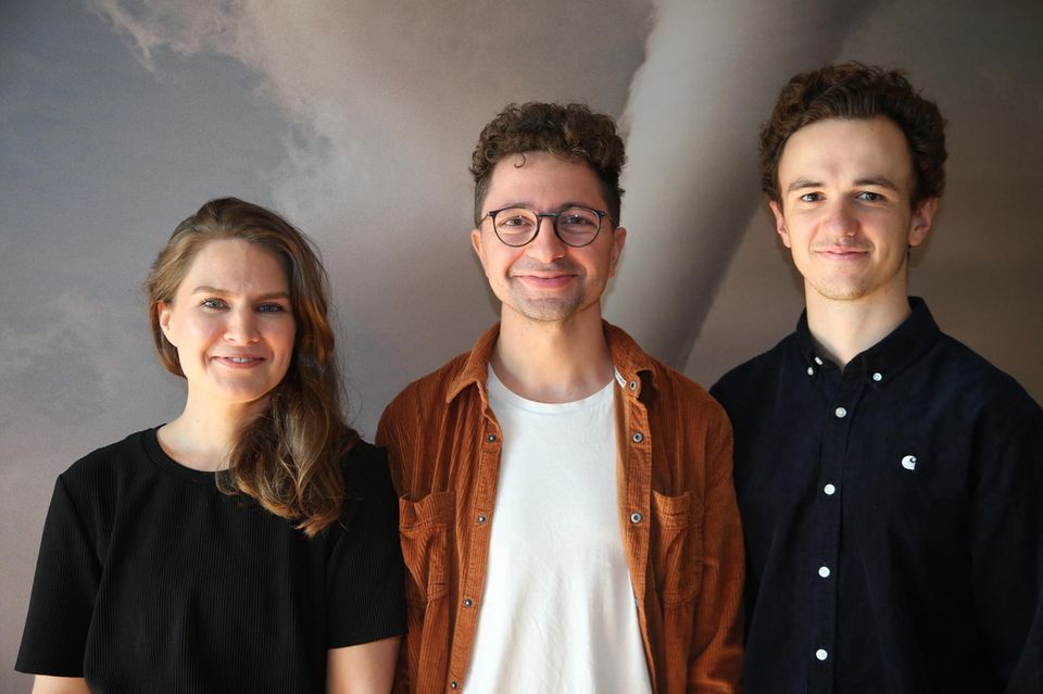 eine junge Frau und zwei junge Männer vor Plakat mit Tornado