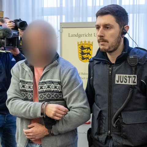 Ein Justizwachtmeister führt im Landgericht einem Mann an Handschellen gefesselt in den Sitzungssaal