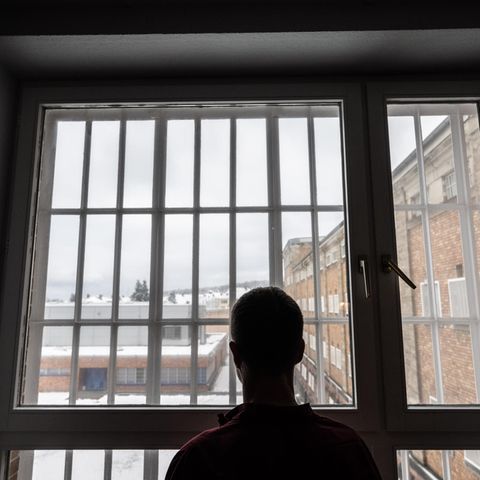 Die Silhouette eines Häftlings ist vor dem vergitterten Fenster einer JVA zu sehen