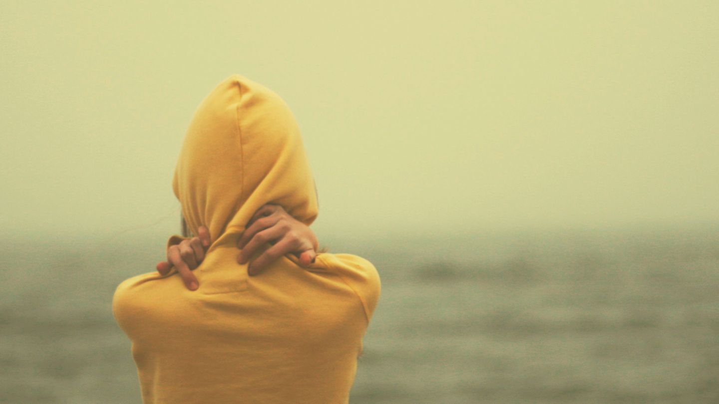Eine Frau steht am Meer und umarmt sich selbst in einem gelben Pullover