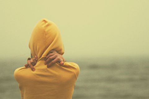 Eine Frau steht am Meer und umarmt sich selbst in einem gelben Pullover