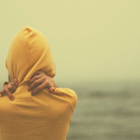 Eine Frau steht am Meer und umarmt sich selbst in einem gelben Pullover