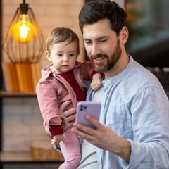 Geldanlage: Ein Mann mit einem Kind auf dem Arm und Smartphone in der Hand