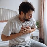 Ein Mann sitzt auf einem Bett und fasst sich an die Brust – offenbar wegen eines Herzinfarktes