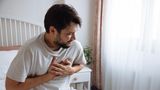 Ein Mann sitzt auf einem Bett und fasst sich an die Brust – offenbar wegen eines Herzinfarktes