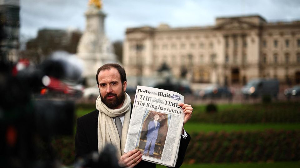 Das Schicksal des Königs hält die Nation in Atem: Ein Reporter berichtet die Lage vor dem Buckingham Palace in London. In seiner Hand hält er die Dienstagsausgabe der Times mit der Schlagzeile "Der König hat Krebs".