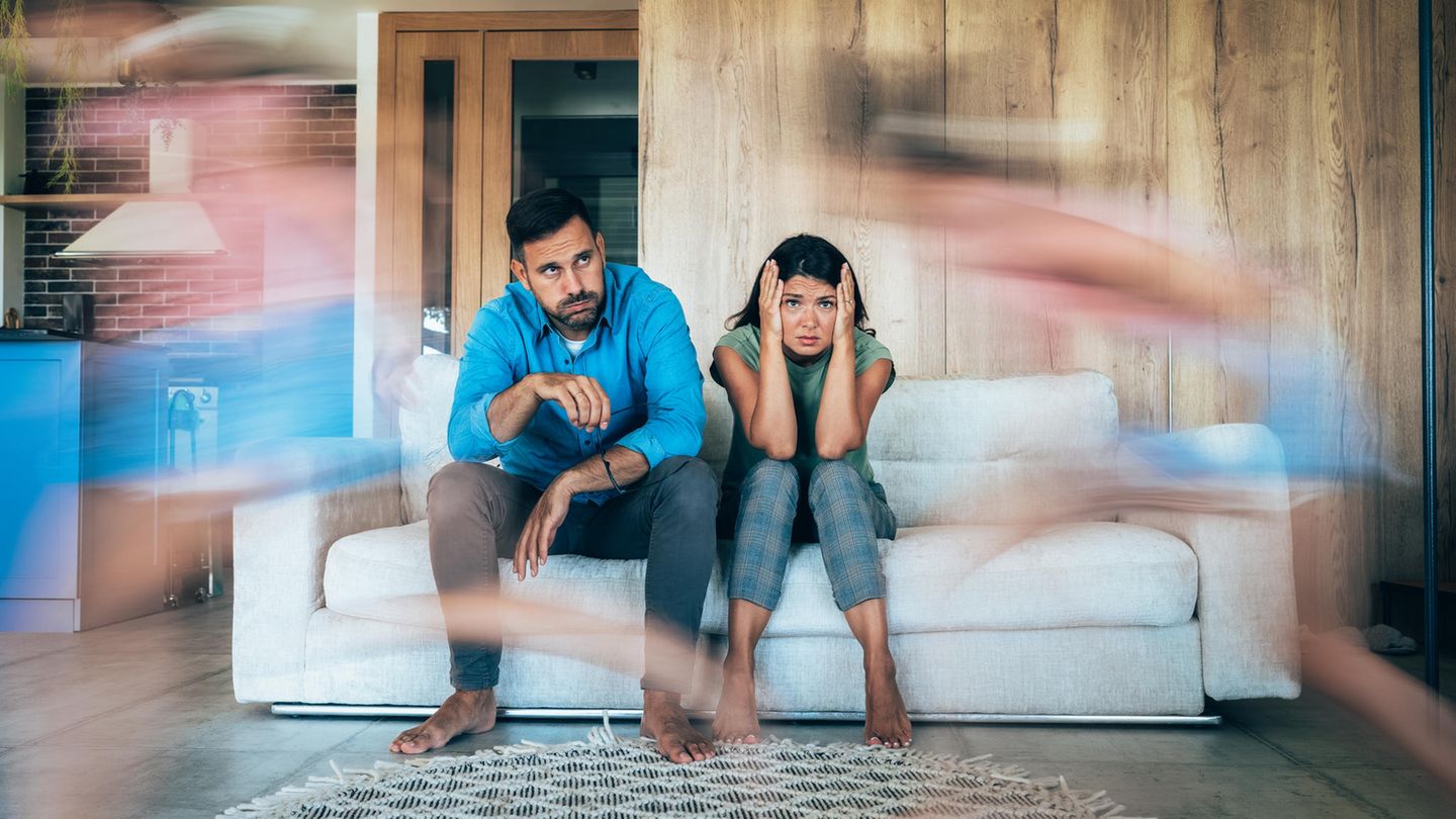 Junge Eltern sitzen erschöpft auf der Couch in Bewegungsunschärfe laufen Kinder um sie herum