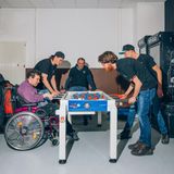 Eine Gruppe Männer, einer im Rollstuhl, spielen Tischfußball und haben Spaß.