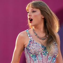 Taylor Swift in einem glitzernden Bühnenanzug steht vor einem pinken Hintergrund und schaut mit geöffnetem Mund zur Seite.
