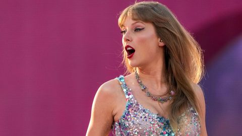 Taylor Swift in einem glitzernden Bühnenanzug steht vor einem pinken Hintergrund und schaut mit geöffnetem Mund zur Seite.