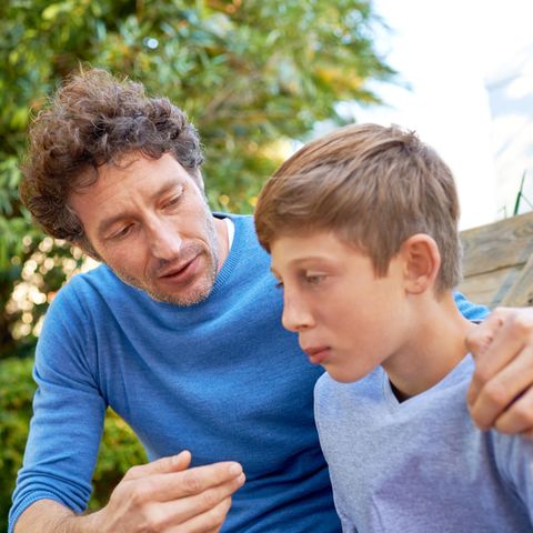 Vater und Sohn sitzen draußen, beide haben einen blauen Pulli an. Der Vater redet auf den Jungen ein