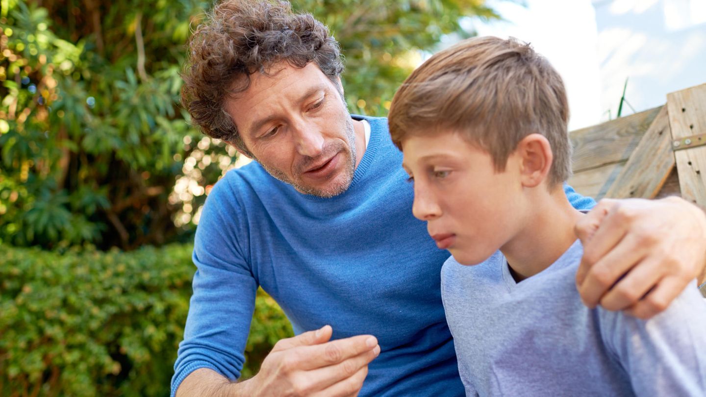 Vater und Sohn sitzen draußen, beide haben einen blauen Pulli an. Der Vater redet auf den Jungen ein