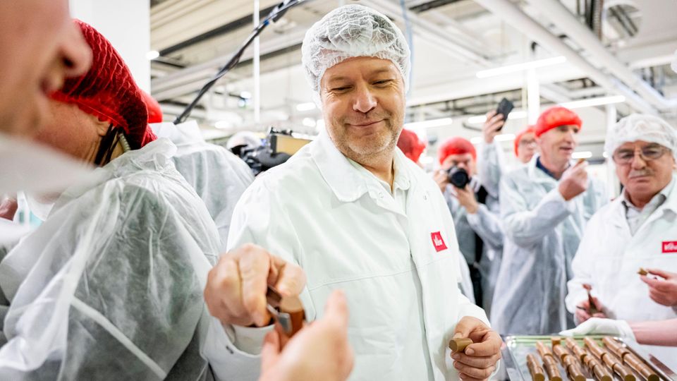 Wirtschaftsminister Robert Habeck, im weißen Kittel und mit Haarnetz, verteilt mit einer Zange Pralinen