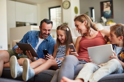 Fröhliche Familie auf dem Sofa surft mit Handys und Tablets im Internet.