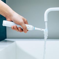 Hand hält eine Zahnbürste unter fließendem Wasser aus Wasserhahn in einem Badezimmer