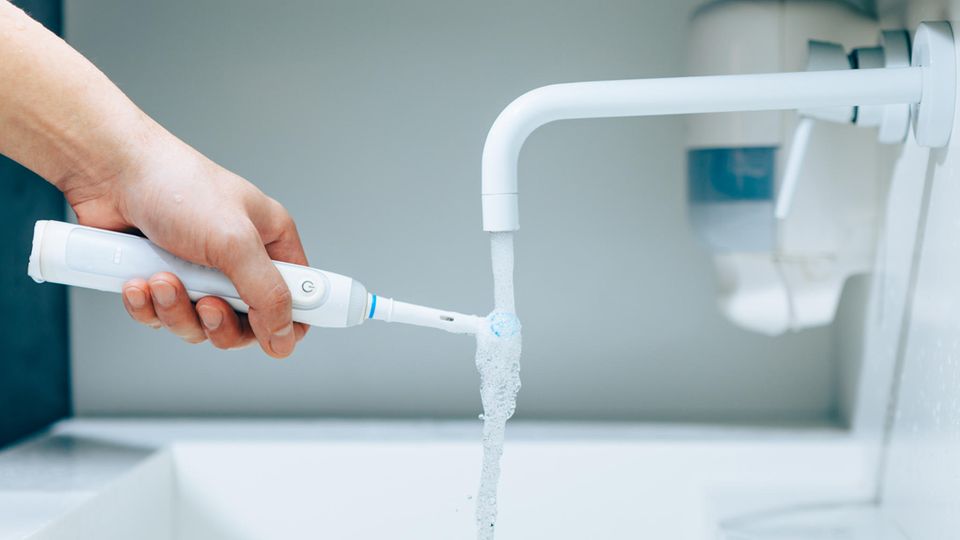 Hand hält eine Zahnbürste unter fließendem Wasser aus Wasserhahn in einem Badezimmer