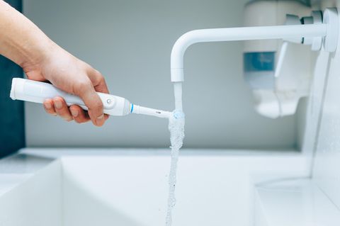 Hand hält eine Zahnbürste unter fließendem Wasser aus Wasserhahn in einem Badezimmer