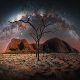 Der Ayers Rock vor einem Sternenhimmel, die Milchstraße zieht einen Bogen über den Horizont