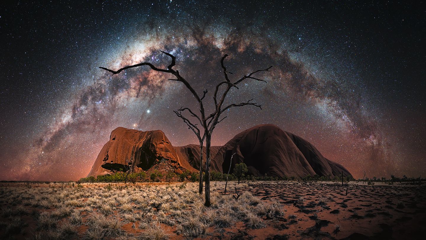 Der Ayers Rock vor einem Sternenhimmel, die Milchstraße zieht einen Bogen über den Horizont