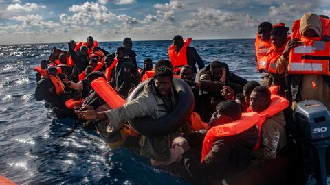 Mehrere Migranten sitzen in einem Boot im Mittelmeer, während Rettungskräfte versuchen ihnen zu helfen