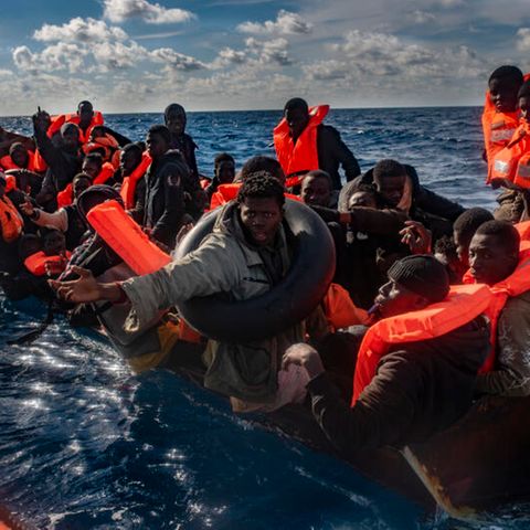 Mehrere Migranten sitzen in einem Boot im Mittelmeer, während Rettungskräfte versuchen ihnen zu helfen