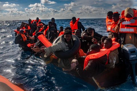 Mehrere Migranten sitzen in einem Boot im Mittelmeer, während Rettungskräfte versuchen ihnen zu helfen