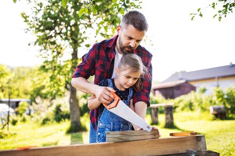 Gartenmöbel selber bauen: Vater und Tochter arbeiten im Garten gemeinsam mit einer Säge.