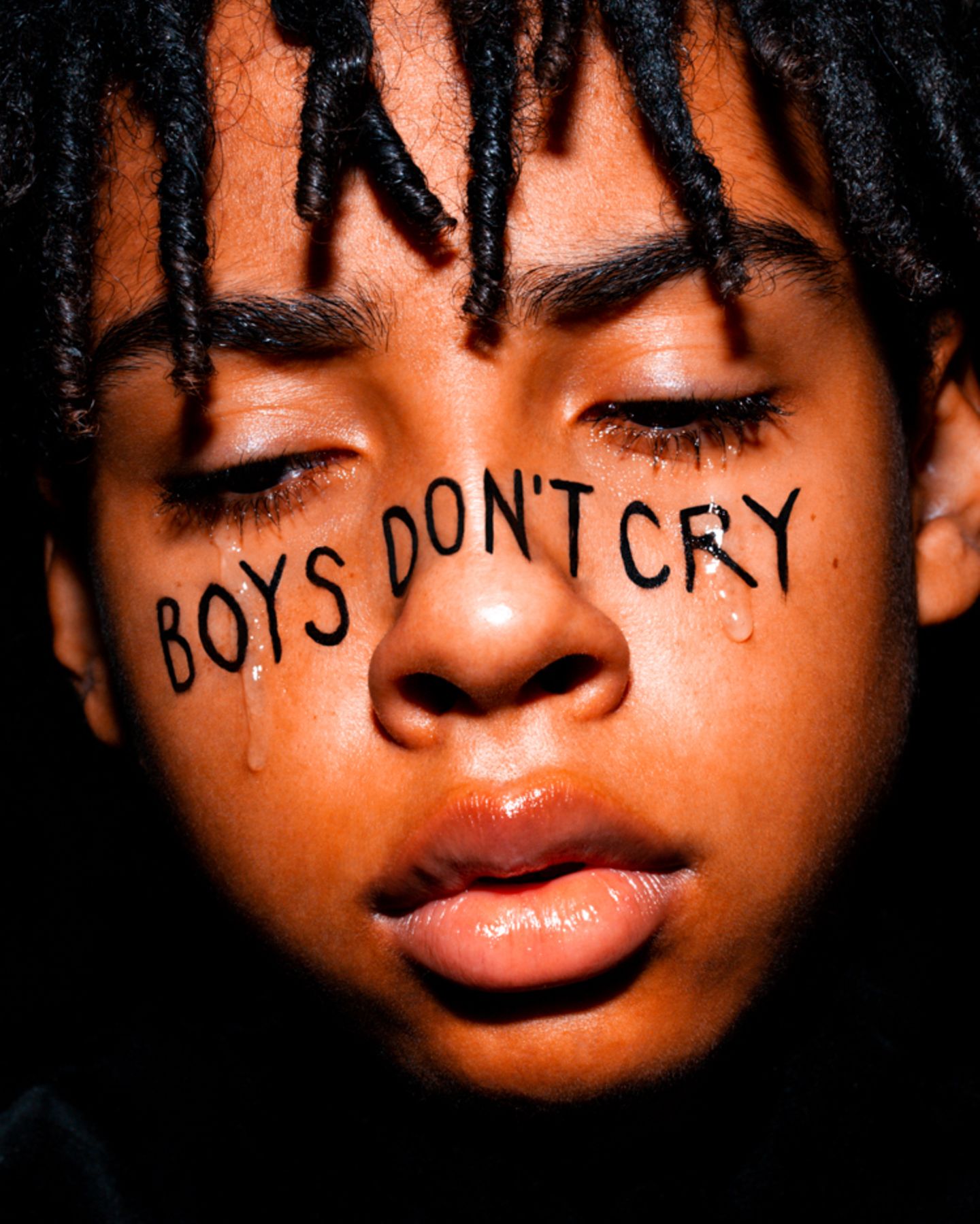 Nahaufnahme Jungengesicht umrahmt von Dreadlocks. Über Wange und Nase in Großbuchstaben in schwarzer Schrift "BOYS DON'T CRY".