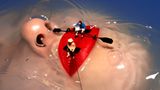 Frauengesicht unter Wasser, nur Nase und roter Mund ragen heraus. Miniaturfiguren sitzen wie Ruderer und Angler auf den Lippen.
