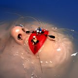 Frauengesicht unter Wasser, nur Nase und roter Mund ragen heraus. Miniaturfiguren sitzen wie Ruderer und Angler auf den Lippen.