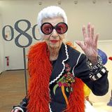 Iris Apfel winkend mit großer Brille und roter Federboa