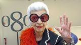 Iris Apfel winkend mit großer Brille und roter Federboa