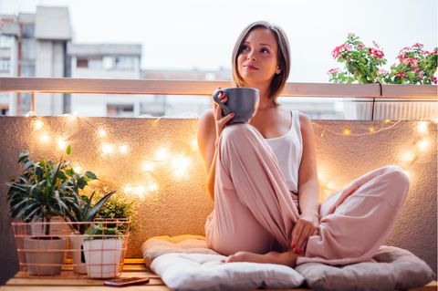 Frau sitzt mit einer Tasse in der Hand auf einem Balkon, der stimmungsvoll beleuchtet ist.