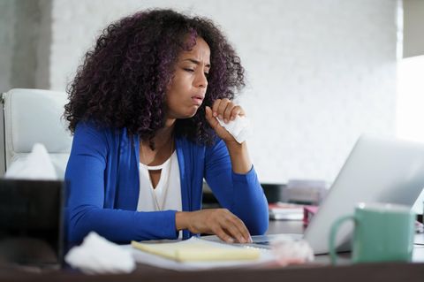 Husten: Junge Frau sitzt im Büro am Laptop und hustet in die Hand